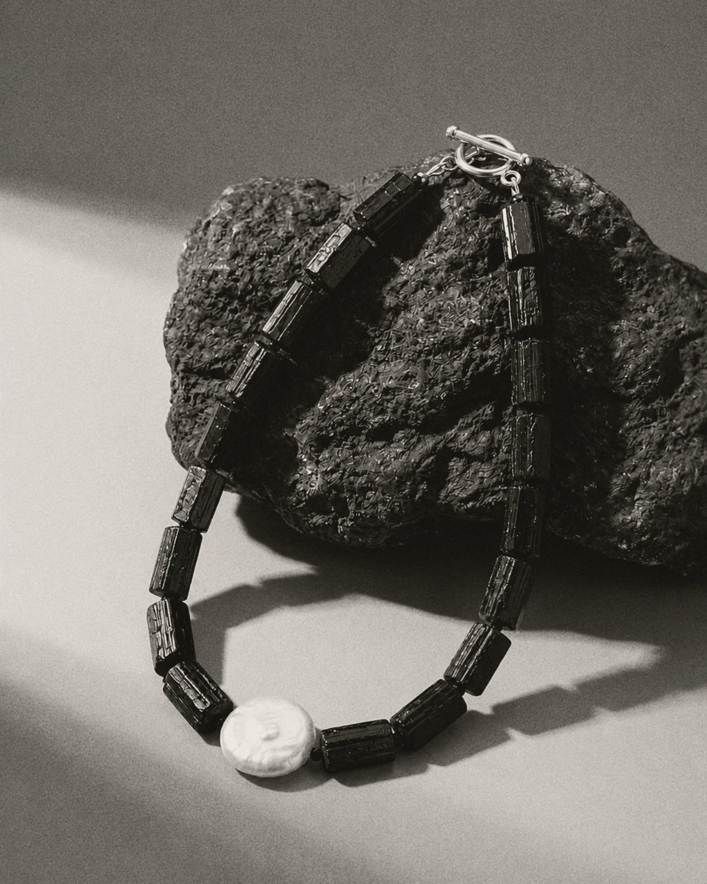 A black beaded necklace with rectangular dark beads and a white oval pendant, draped over a rough dark rock on a light gray surface.