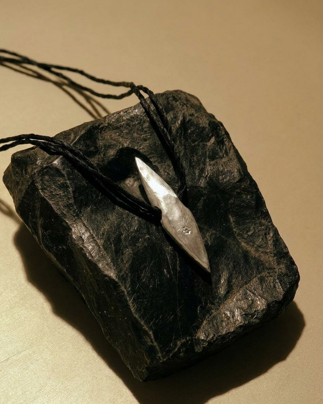 A silver pointed pendant necklace on a black cord, displayed on a dark rough stone against a warm beige background.