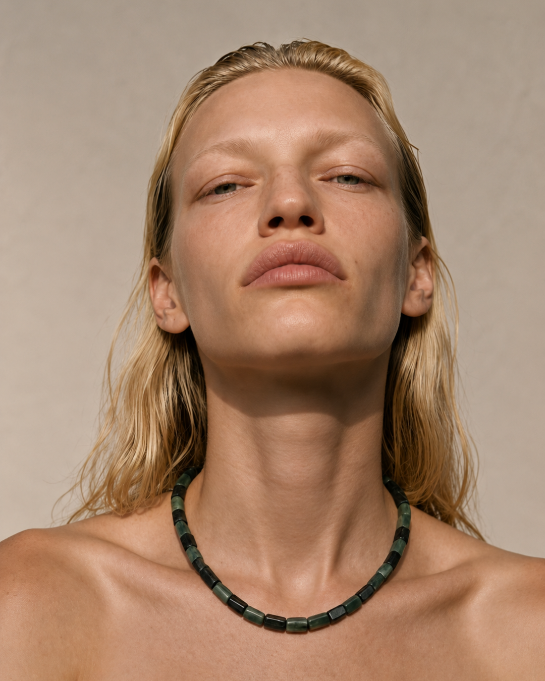 A close-up portrait of a woman with blonde hair wearing a dark green beaded necklace against a neutral beige background.