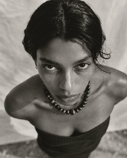 Black and white close-up portrait of a woman looking directly at the camera, wearing a dark beaded necklace and a dark strapless top against a light draped background.