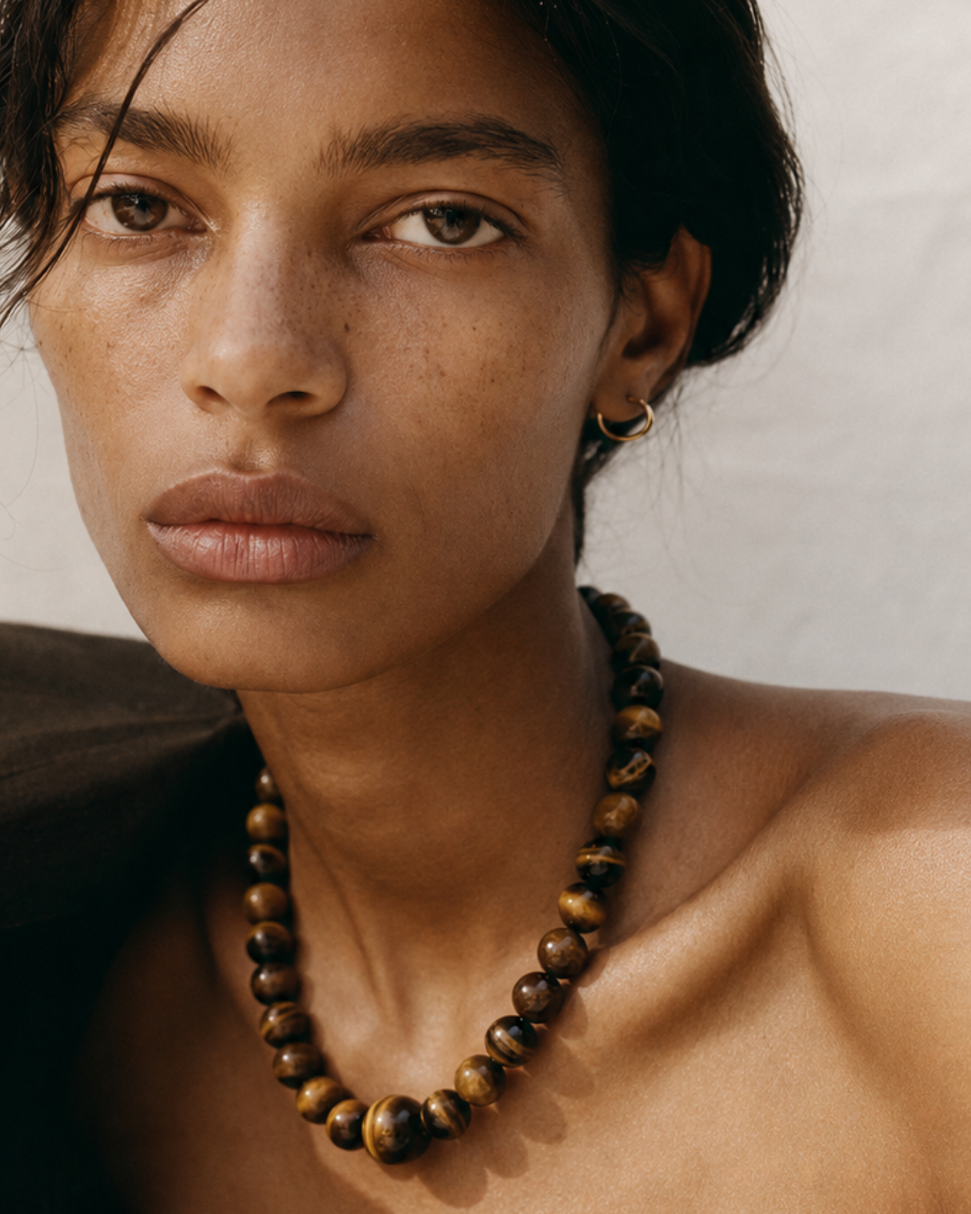 A close-up portrait of a woman wearing a chunky beaded necklace made of brown and gold tiger's eye beads, with a small gold hoop earring, against a neutral light background.