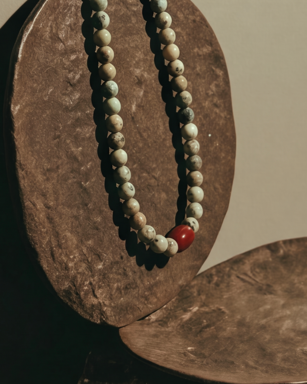 A beaded necklace with light beige and gray marbled beads and a single red accent bead, draped over a textured brown wooden display stand.