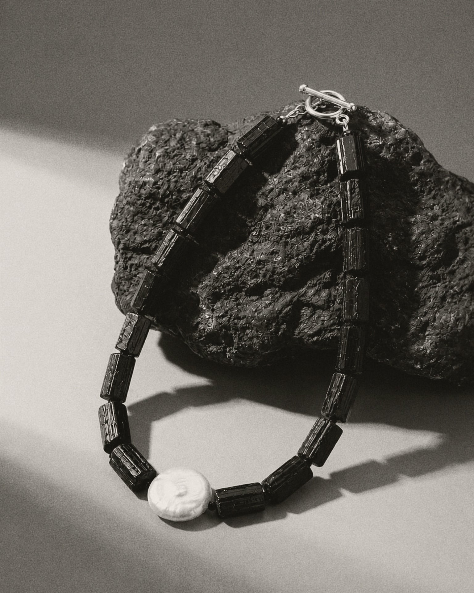 A black beaded necklace with rectangular dark beads and a white oval pendant, draped over a rough dark rock on a light gray surface.