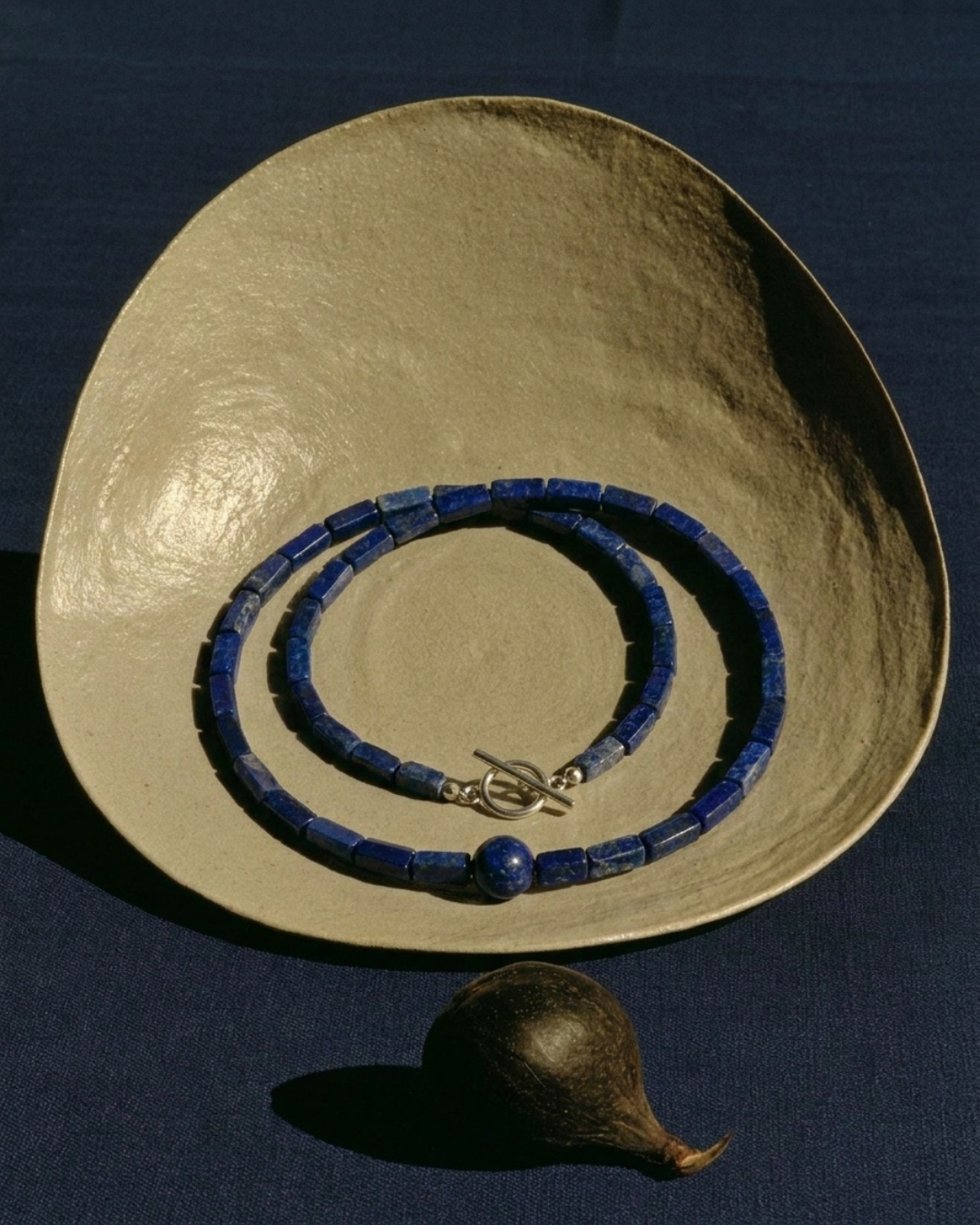 A beaded necklace made of dark blue cylindrical beads with a silver toggle clasp and a single round blue bead, displayed on a large textured beige plate over a dark blue background.