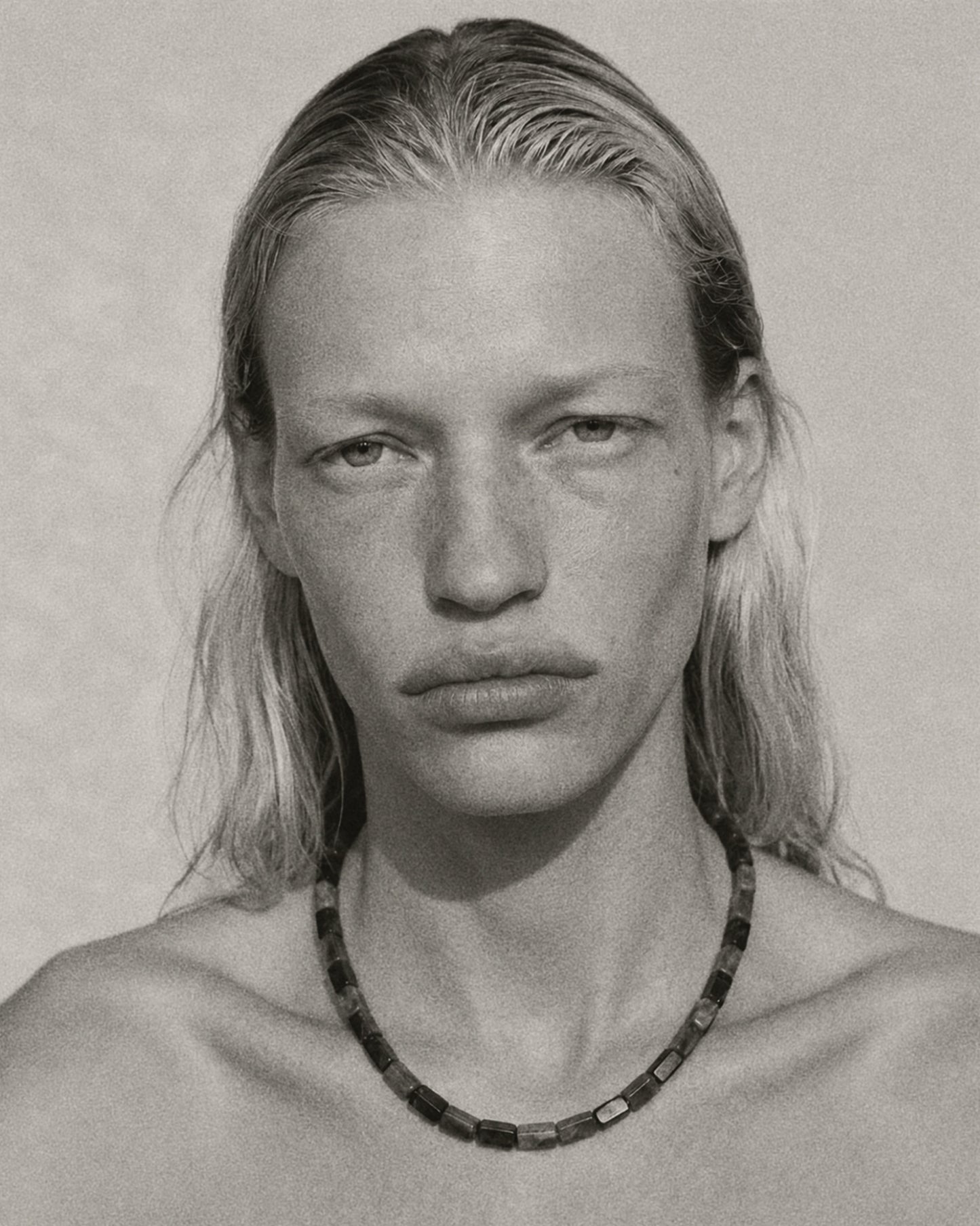 A black and white close-up portrait of a person with long hair wearing a dark beaded necklace against a plain light background.