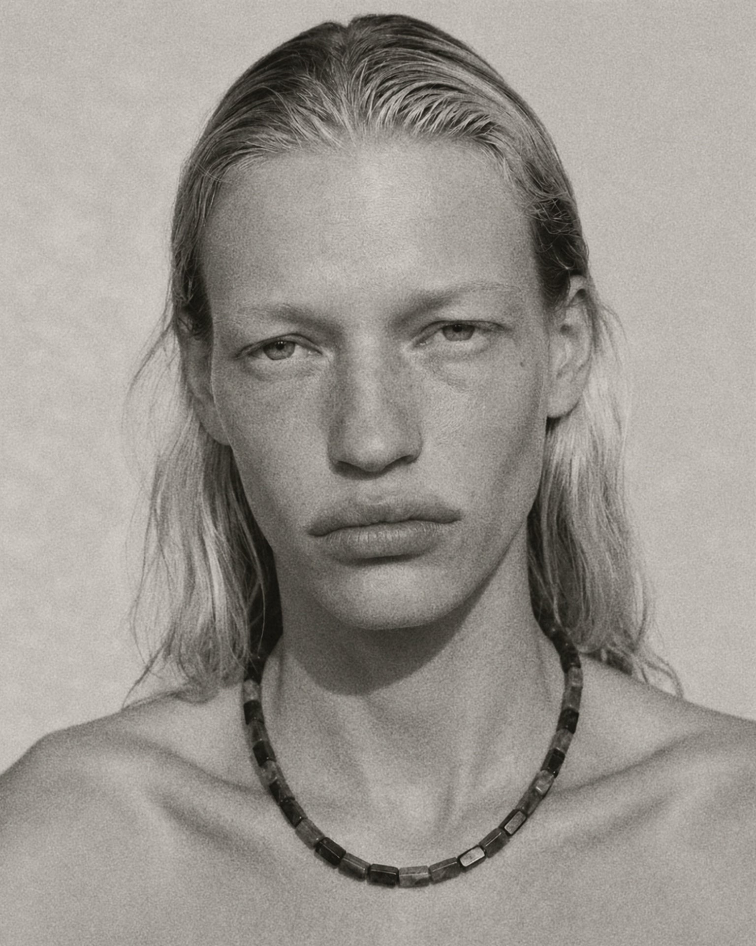 A black and white close-up portrait of a person with long hair wearing a dark beaded necklace against a plain light background.
