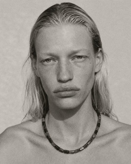 A black and white close-up portrait of a person with long hair wearing a dark beaded necklace against a plain light background.