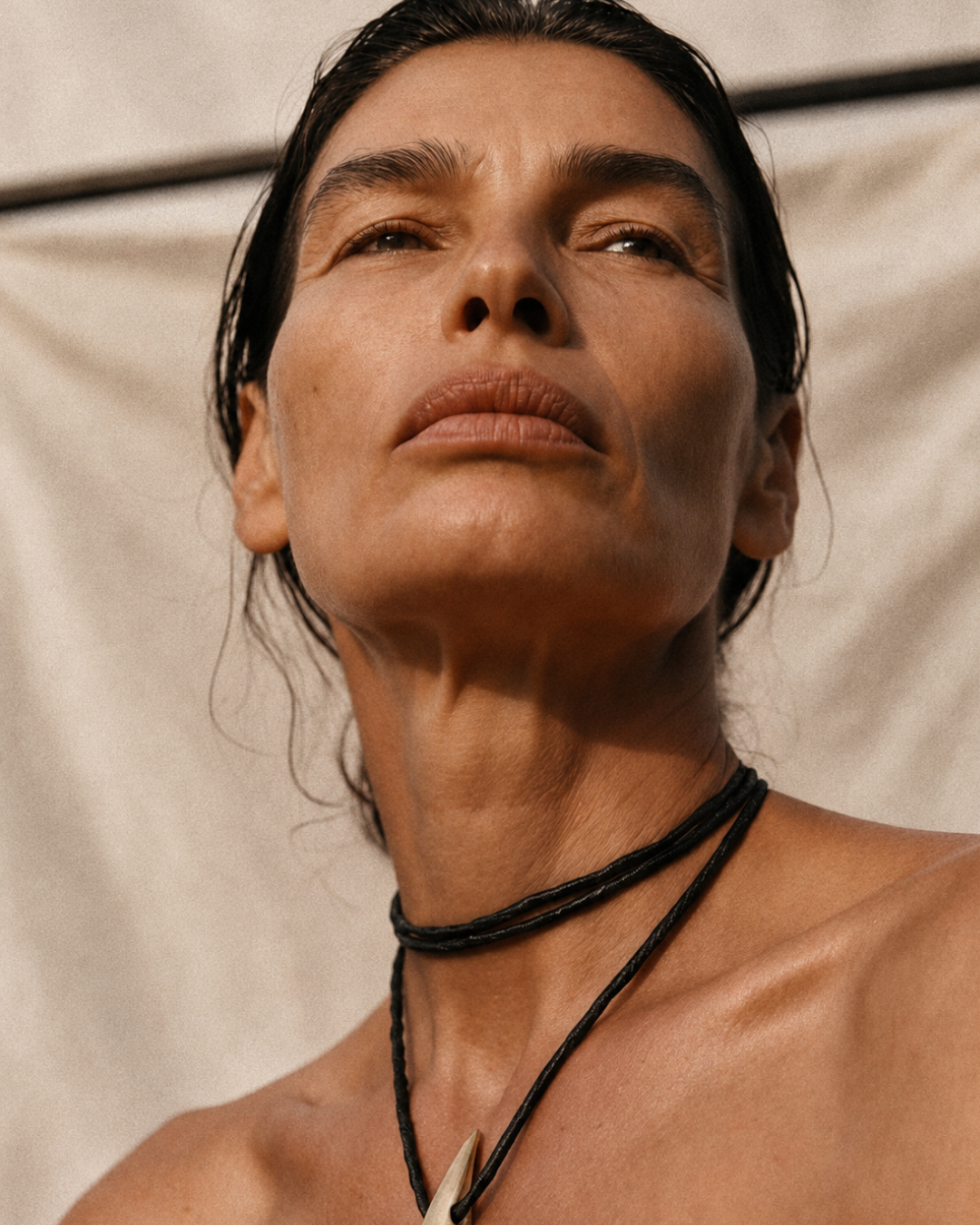 A close-up portrait of a woman wearing a black multi-strand cord choker necklace and a black cord necklace with a white pointed pendant, set against a neutral beige fabric background.