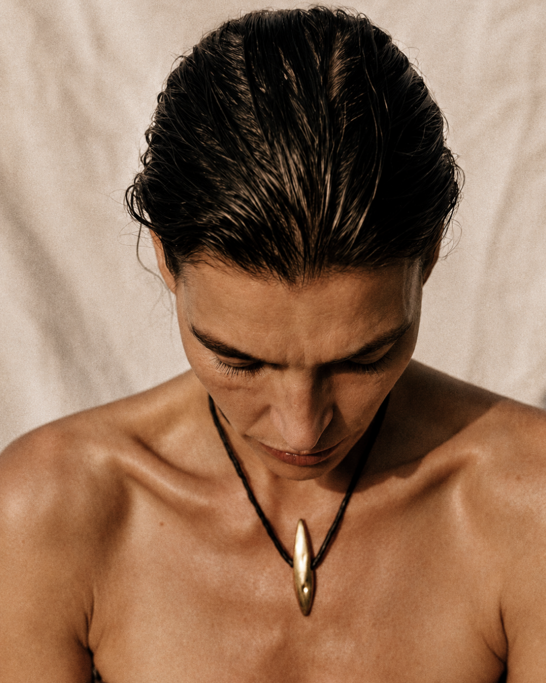 A close-up of a person with dark hair looking down, wearing a black cord necklace with a polished gold pendant against a neutral beige fabric background.