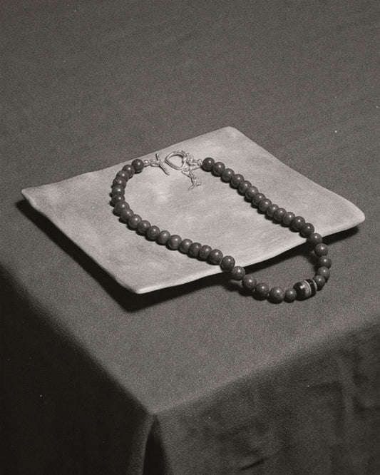 A beaded necklace with a silver-toned toggle clasp and small pendant, draped over a square fabric cushion on a dark draped surface.