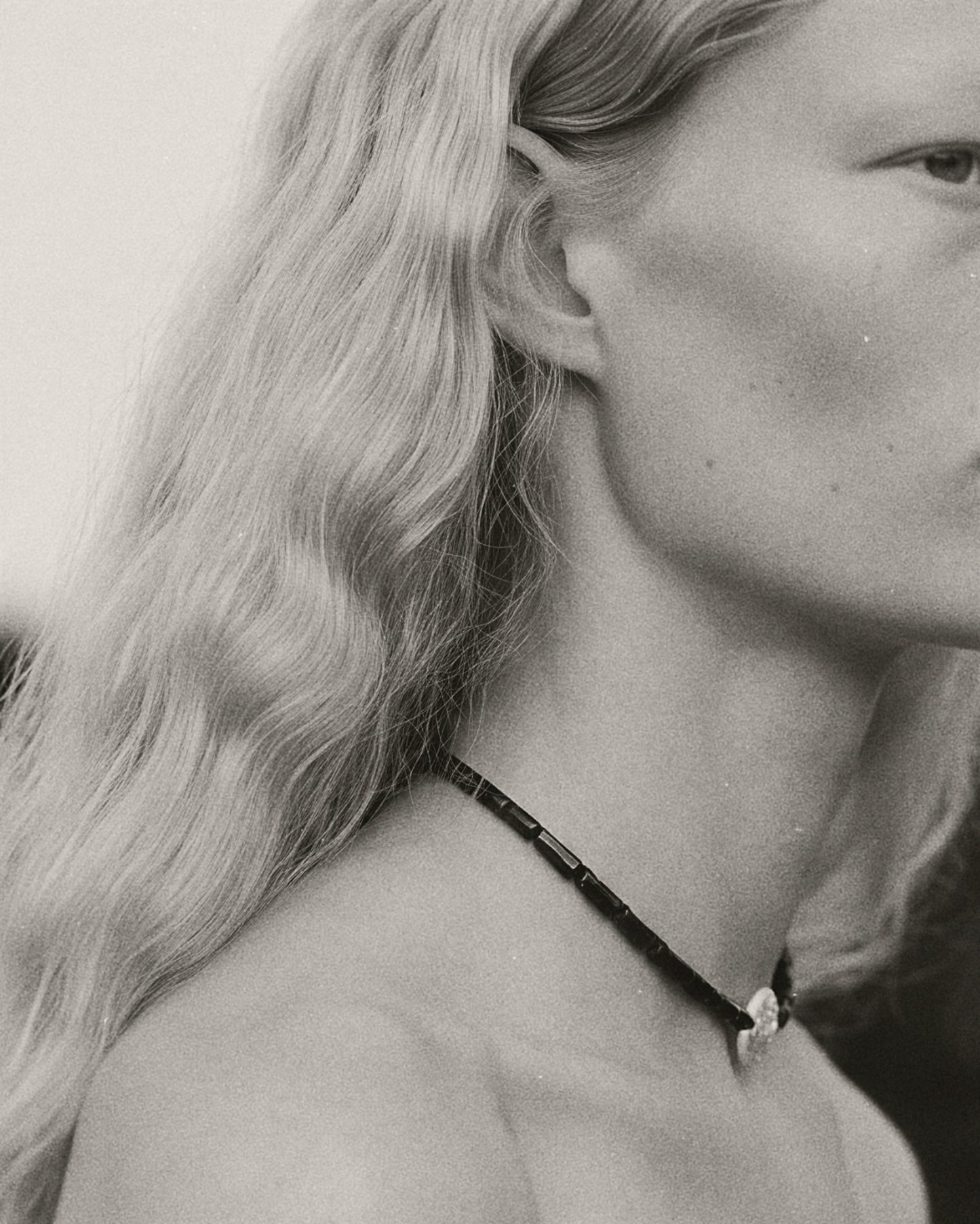 A close-up side profile of a woman with long wavy blonde hair wearing a dark beaded necklace with a small silver-toned pendant.