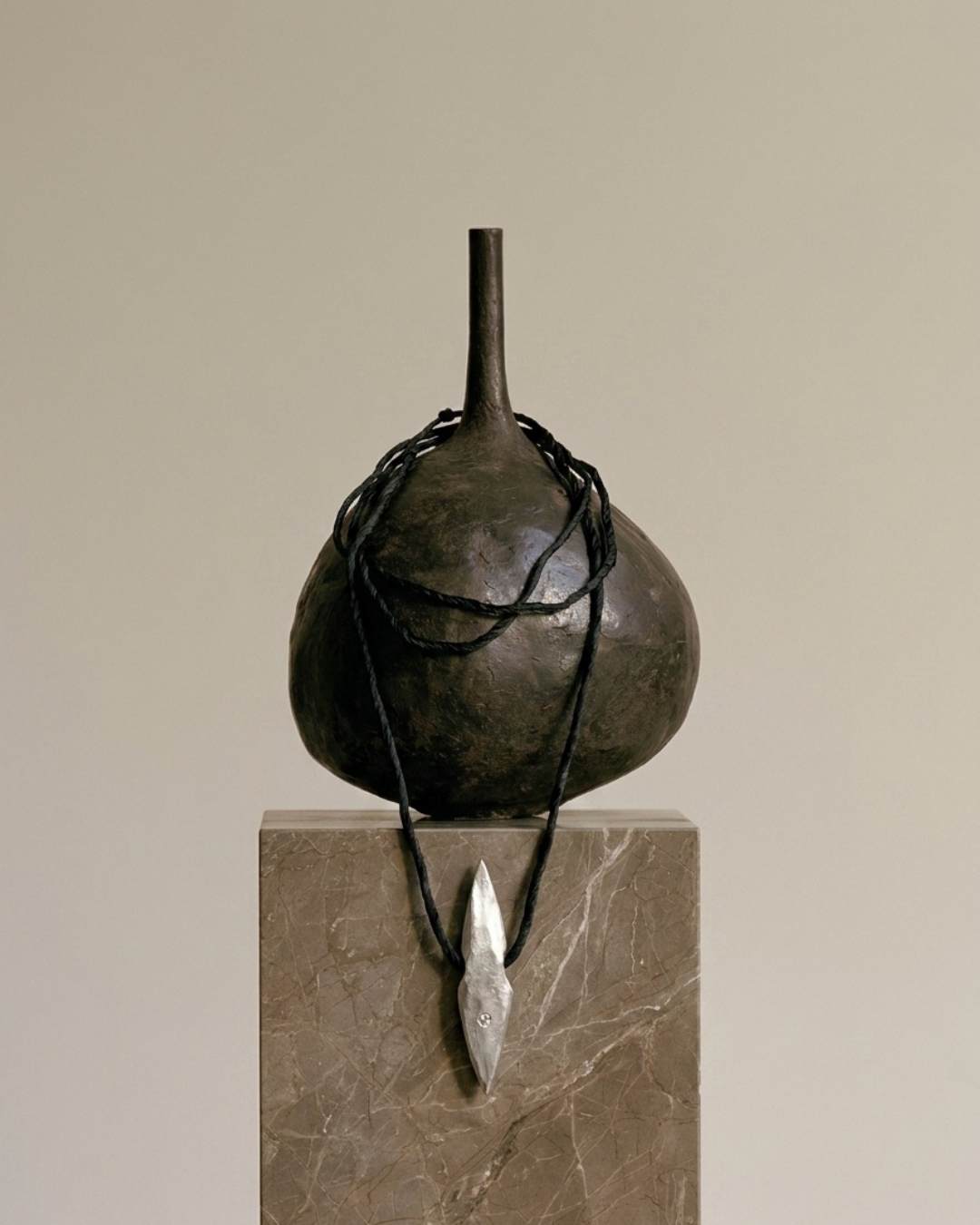 A dark metallic sculpture resembling a rounded bulb with a narrow neck, wrapped with black cords and hanging from a stone pedestal, set against a plain beige background.