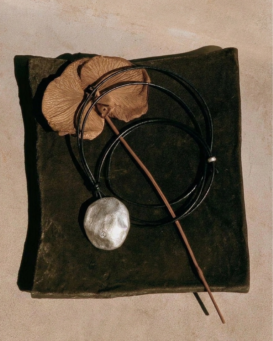 A rustic pendant necklace featuring a textured silver stone on a thin brown cord, styled with dried brown leaves on a dark square plate over a beige background.