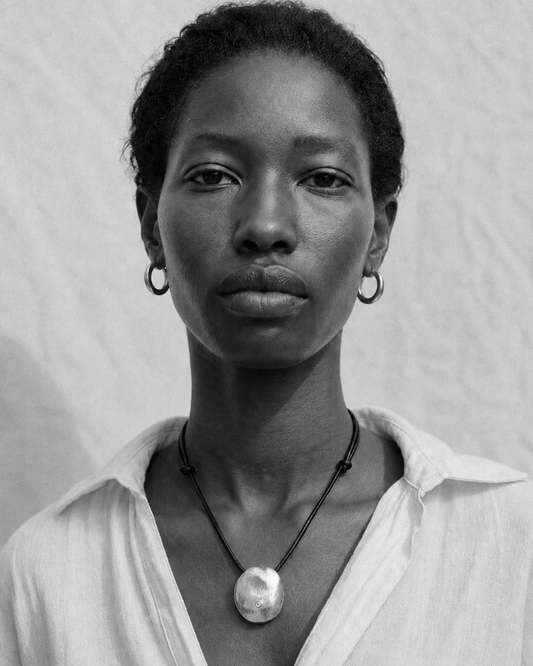 A black and white portrait of a woman wearing a dark cord necklace with a large, textured, silver-toned oval pendant. She is also wearing simple hoop earrings and a light-colored collared shirt, set against a neutral, textured background.