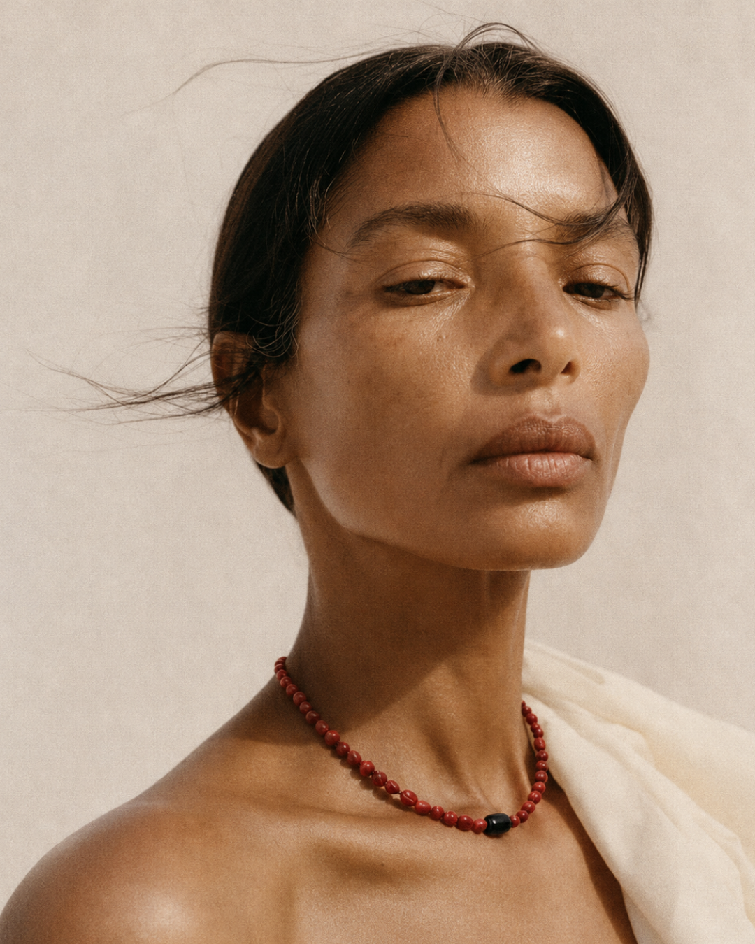A close-up portrait of a woman wearing a red beaded necklace with a single dark accent bead, set against a warm beige background with a white draped fabric.