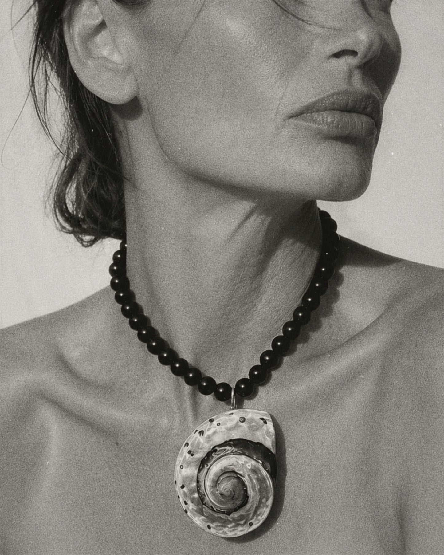 A close-up black and white photograph of a model wearing a dark beaded necklace with a large spiral seashell pendant.