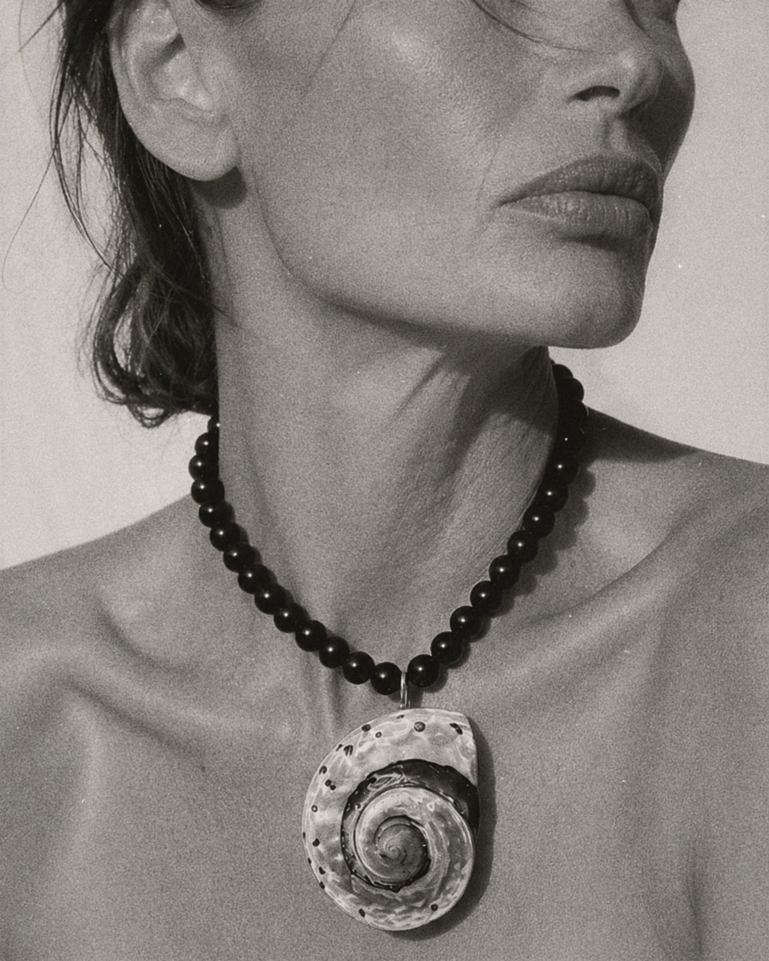 A close-up black and white photograph of a model wearing a dark beaded necklace with a large spiral seashell pendant.