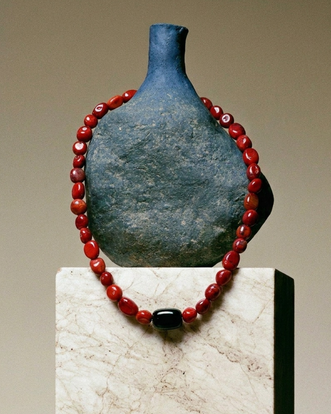 A red beaded necklace with a glossy black central bead, displayed around a dark blue ceramic vase on a light marble pedestal against a beige background.