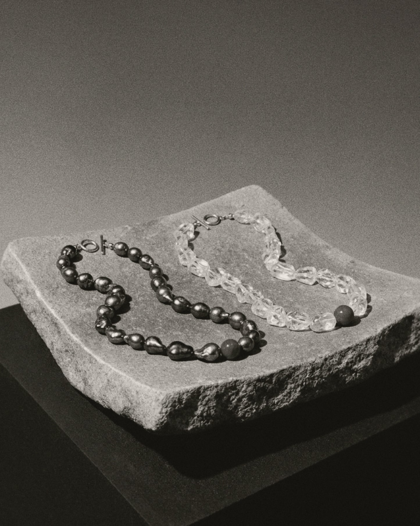 Two beaded necklaces displayed on a rough stone slab against a dark gray studio background. One necklace has dark, glossy round beads, and the other has translucent clear textured beads.