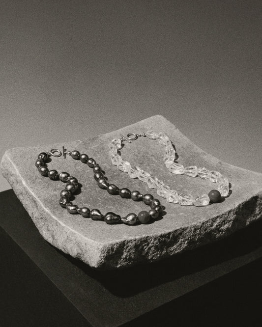 Two beaded necklaces displayed on a rough stone slab against a dark gray studio background. One necklace has dark, glossy round beads, and the other has translucent clear textured beads.