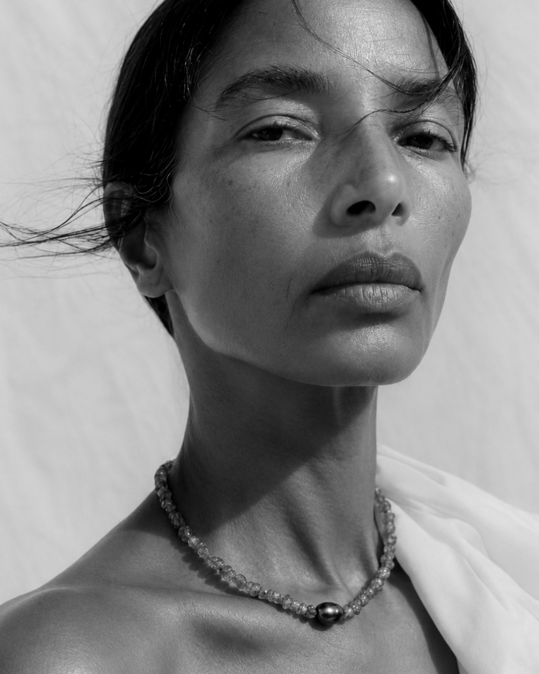 A close-up black and white portrait of a woman wearing a delicate beaded necklace with a central dark bead, set against a plain textured background.
