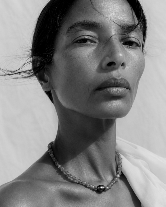 A close-up black and white portrait of a woman wearing a delicate beaded necklace with a central dark bead, set against a plain textured background.