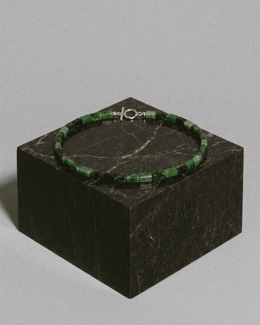 A beaded bracelet made of green faceted beads with a silver-toned clasp, displayed on a dark marble cube against a light gray background.