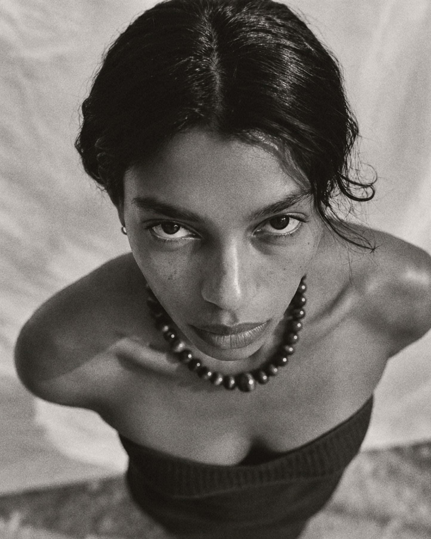 Black and white close-up portrait of a woman looking directly at the camera, wearing a dark beaded necklace and a dark strapless top against a light draped background.