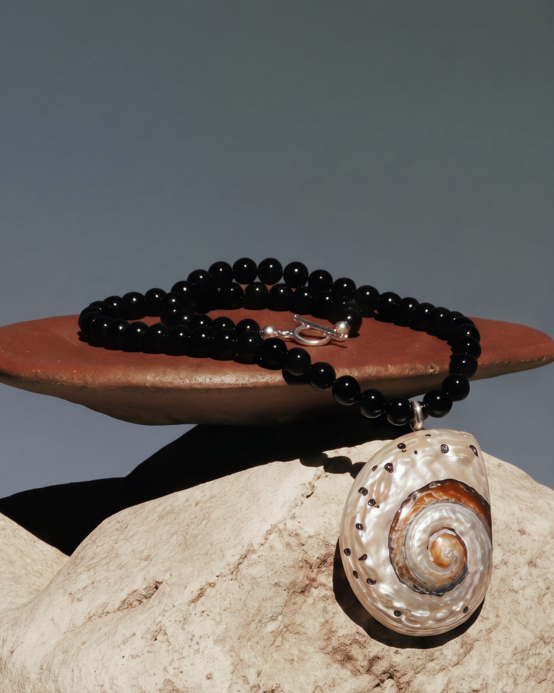 A black beaded necklace with a large spiral shell pendant, displayed on a terracotta dish and stone against a gray background.
