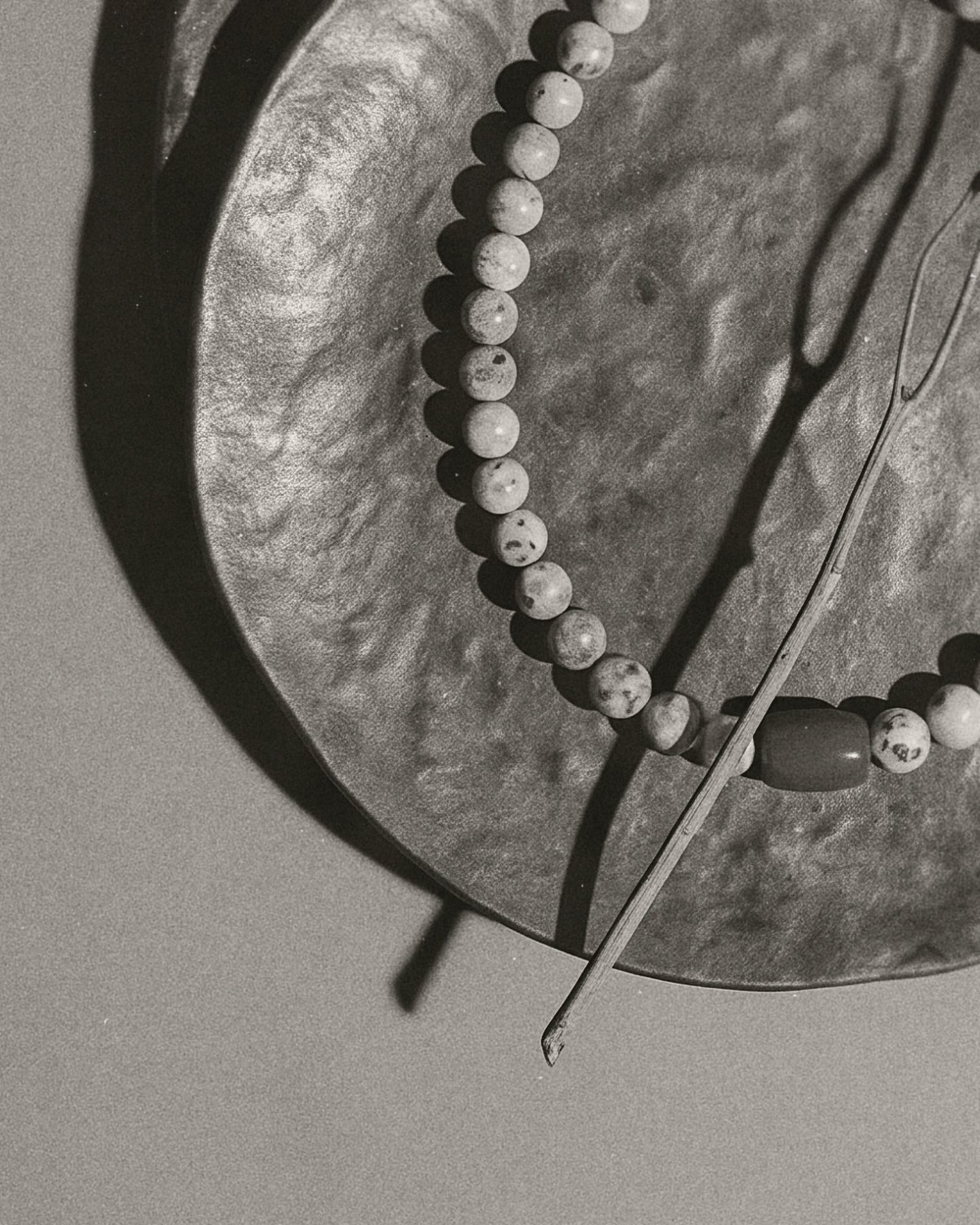A close-up black and white photograph of a beaded necklace draped over a textured circular plate, with a thin twig resting diagonally across the frame.