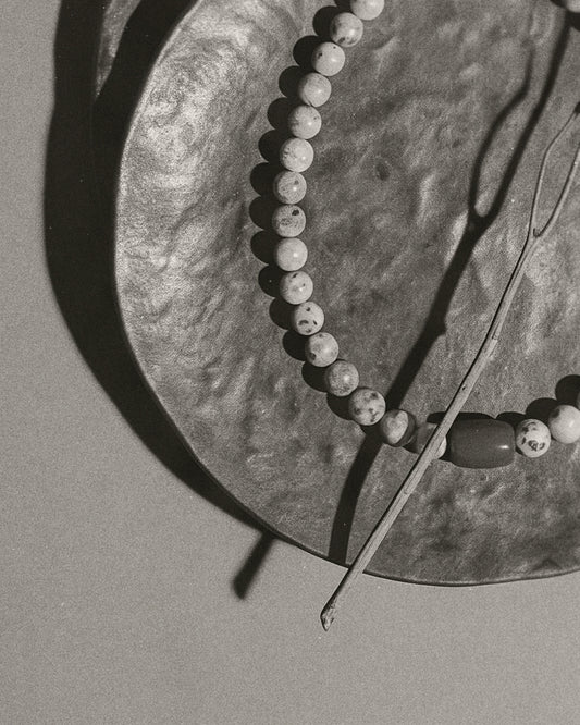 A close-up black and white photograph of a beaded necklace draped over a textured circular plate, with a thin twig resting diagonally across the frame.