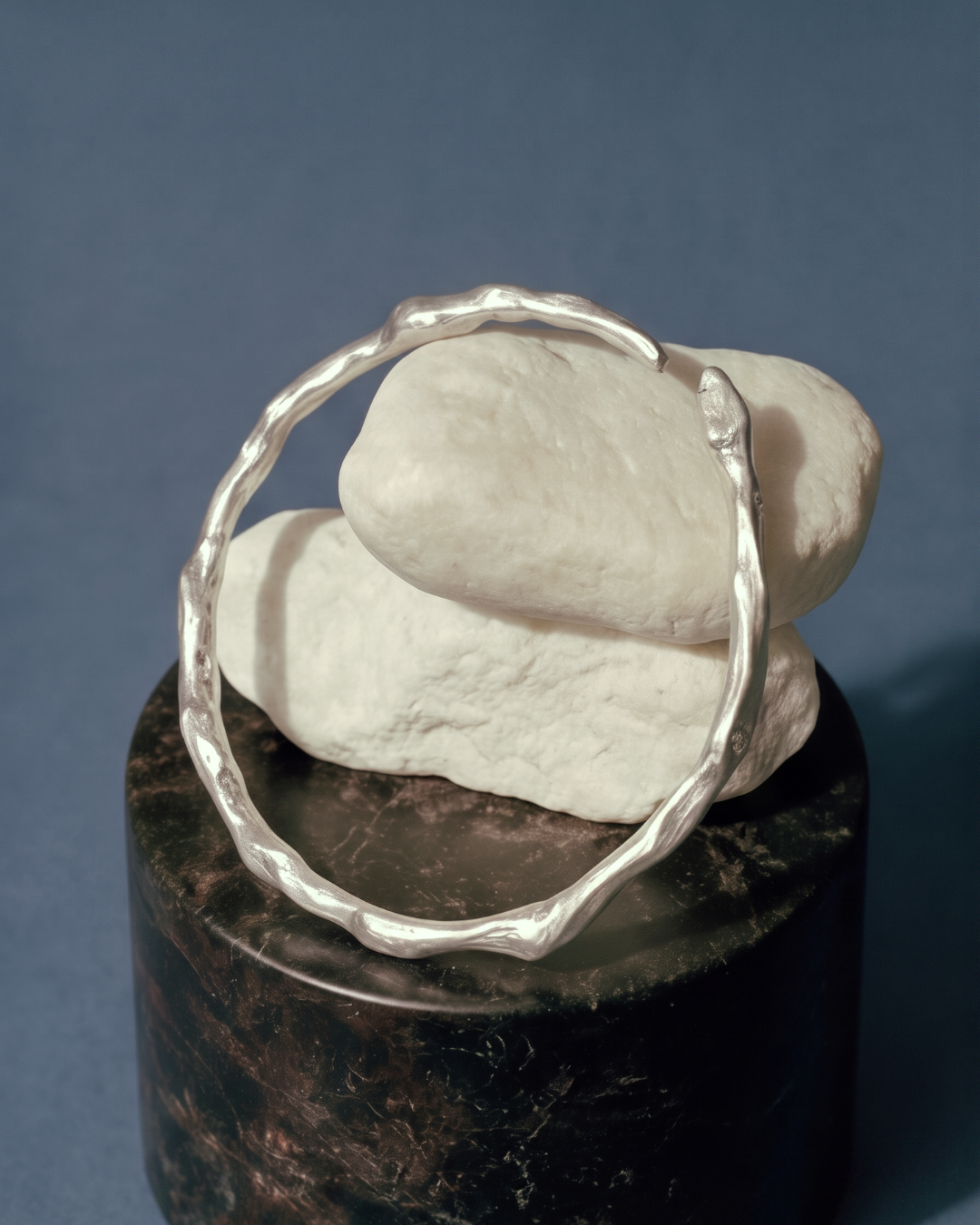 A hammered silver bangle bracelet displayed on stacked white stones atop a dark round pedestal against a blue background.