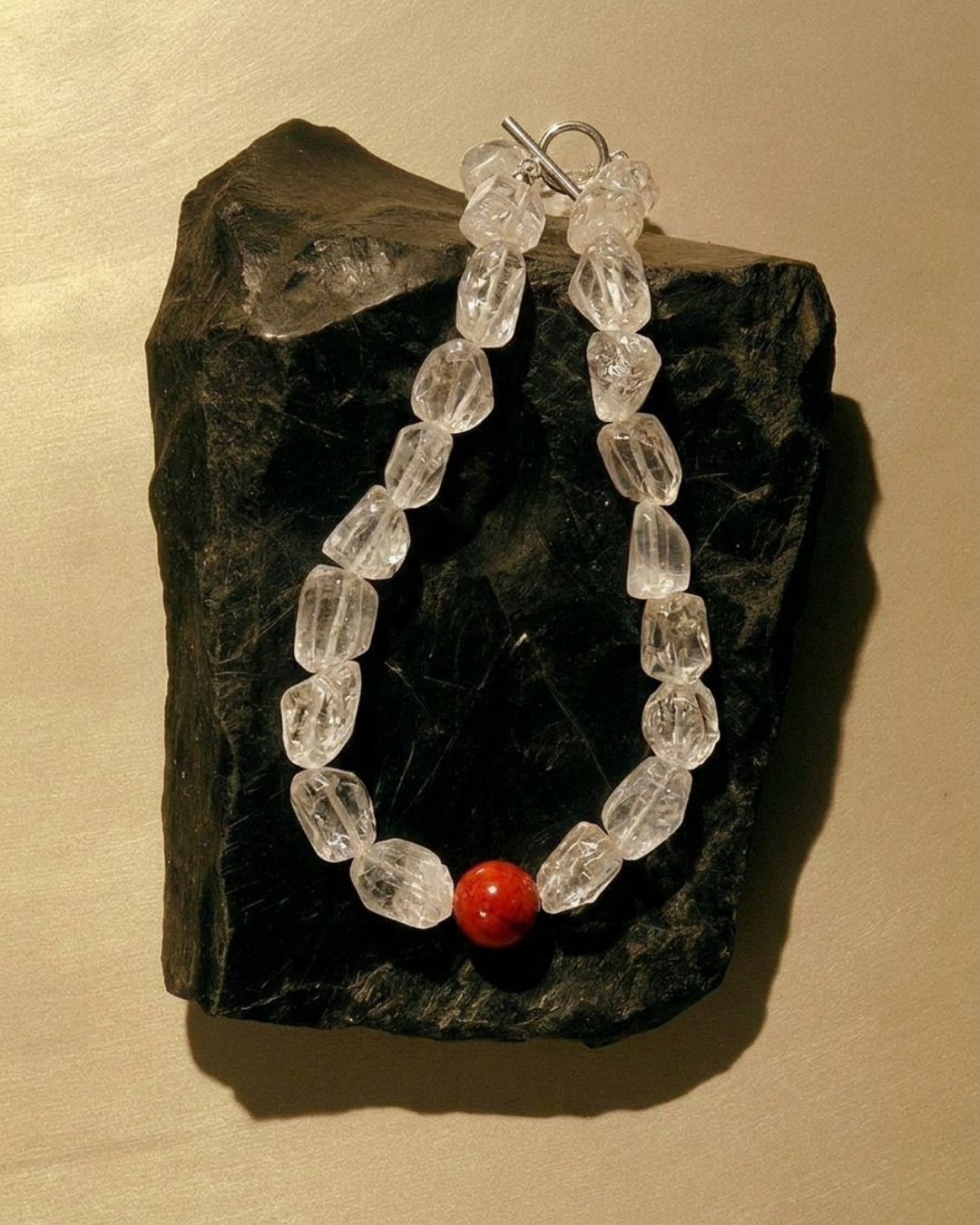 A beaded necklace made of faceted clear crystal-like beads with a single small red bead at the bottom, displayed on a dark rock against a warm beige background.