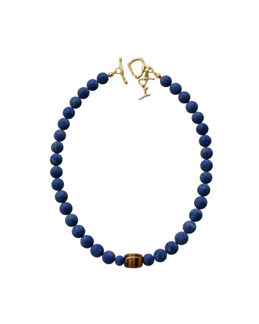 A beaded necklace featuring dark blue lapis lazuli stones and a single brown tiger's eye bead, finished with a gold toggle clasp on a white background.