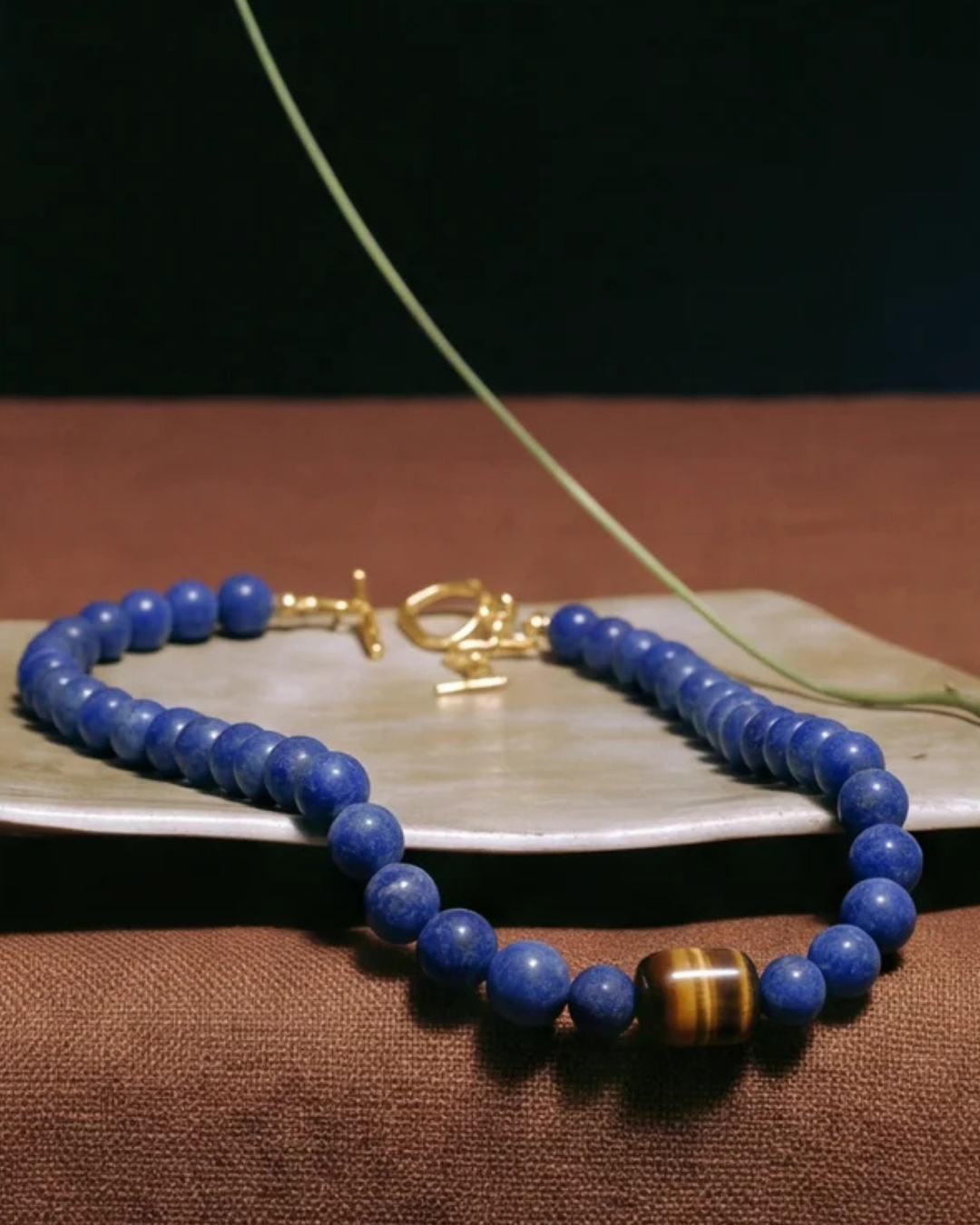 A beaded necklace with round blue beads, a gold clasp, and a tiger's eye accent bead, displayed on a light stone dish over a brown textured surface.