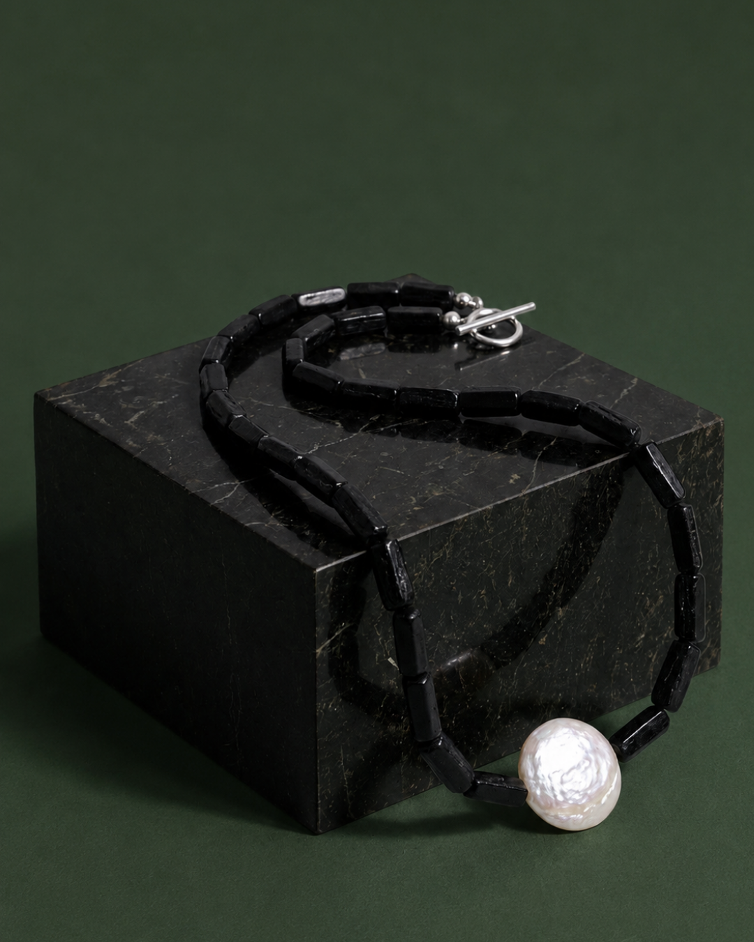 A black beaded necklace with a large white baroque pearl pendant, draped over a black marble display block against a dark green background.
