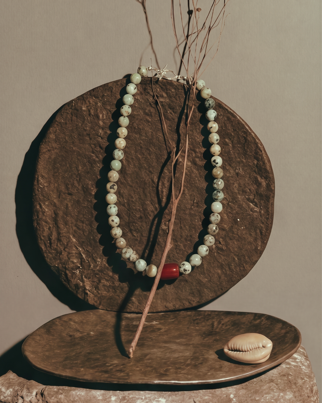A beaded necklace with pale green and white marbled beads and a single red accent bead, draped over a round brown stone display with a small cowrie shell on a metal tray below.