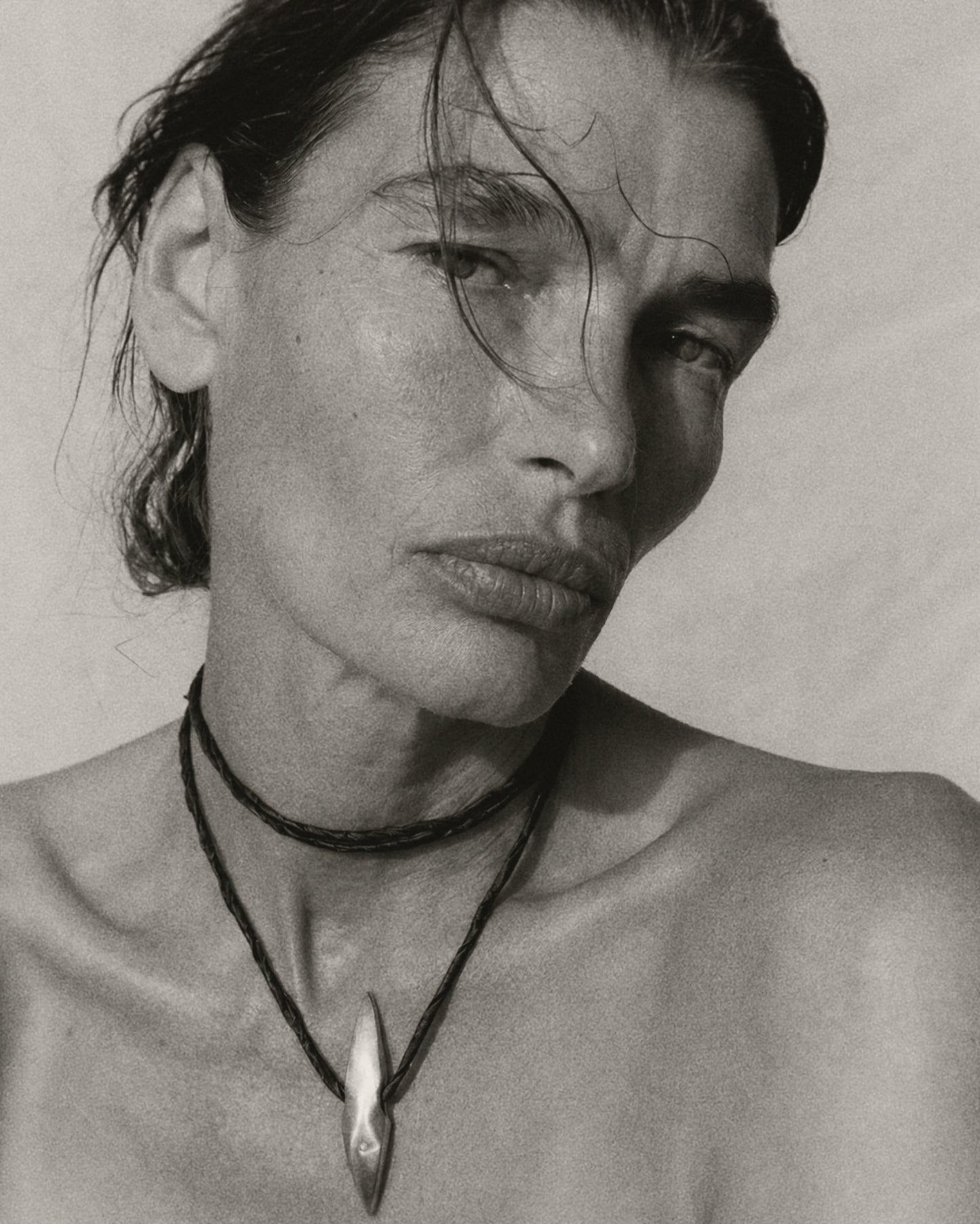 A black and white close-up portrait of a woman wearing layered necklaces, including a prominent pendant necklace with a teardrop-shaped silver pendant.