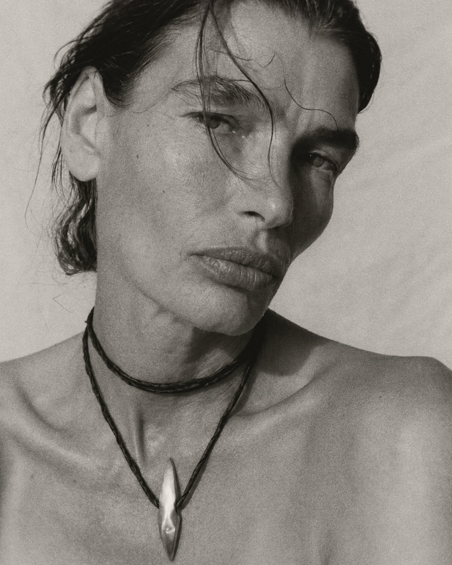 A black and white close-up portrait of a woman wearing layered necklaces, including a prominent pendant necklace with a teardrop-shaped silver pendant.