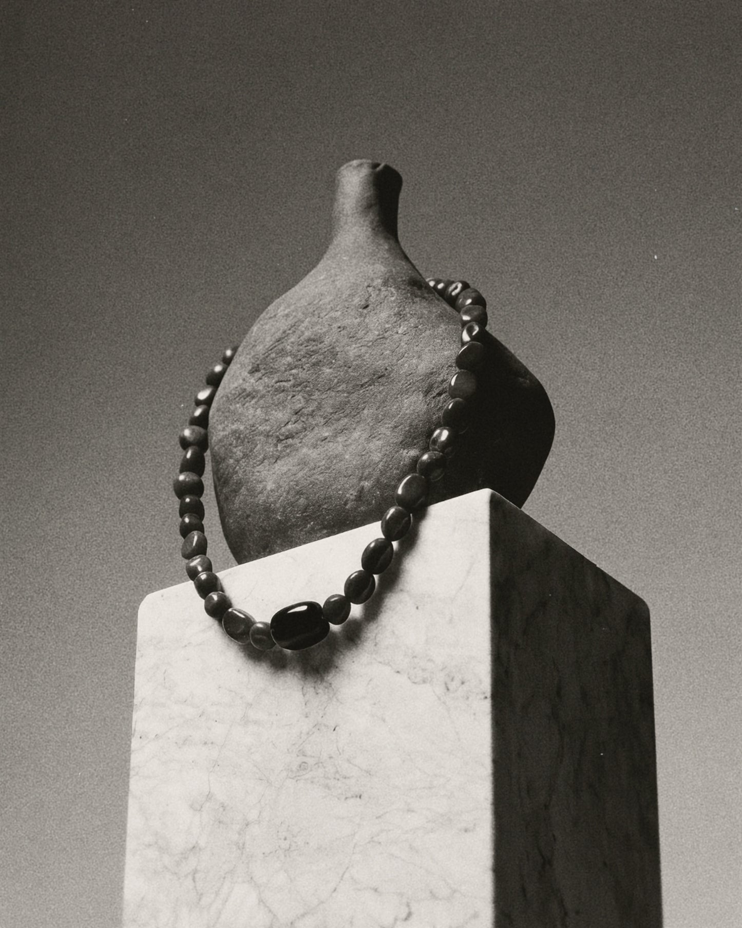 A dark gray ceramic bottle with a short neck, displayed on a white marble pedestal with a dark beaded necklace draped around it, in a black and white photograph.