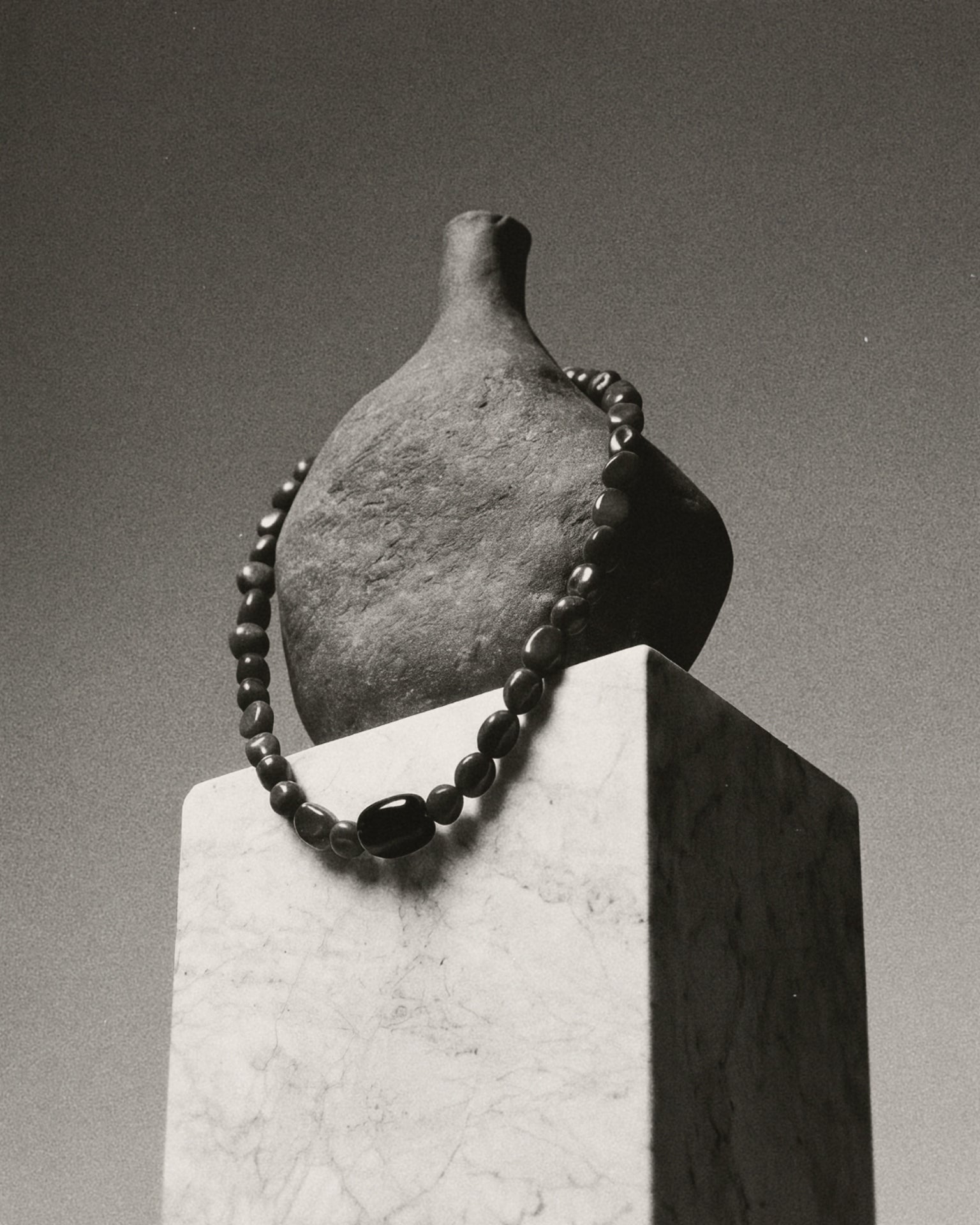 A dark gray ceramic bottle with a short neck, displayed on a white marble pedestal with a dark beaded necklace draped around it, in a black and white photograph.