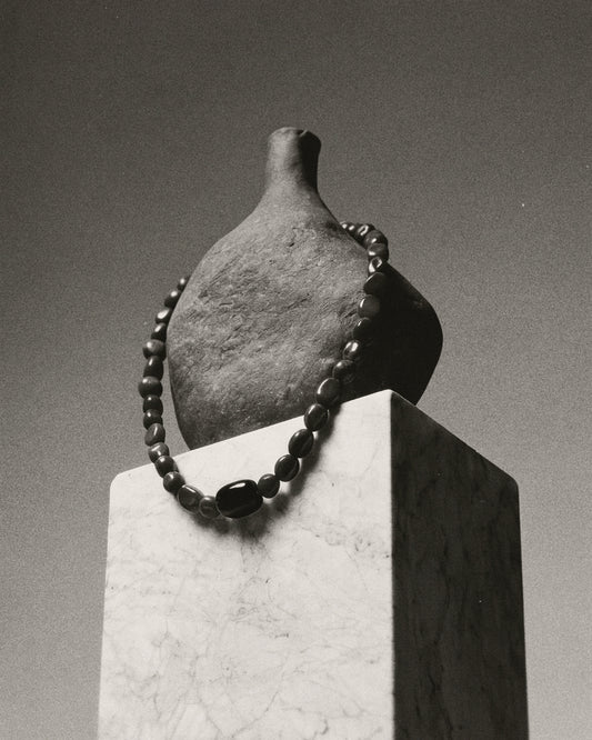 A dark gray ceramic bottle with a short neck, displayed on a white marble pedestal with a dark beaded necklace draped around it, in a black and white photograph.