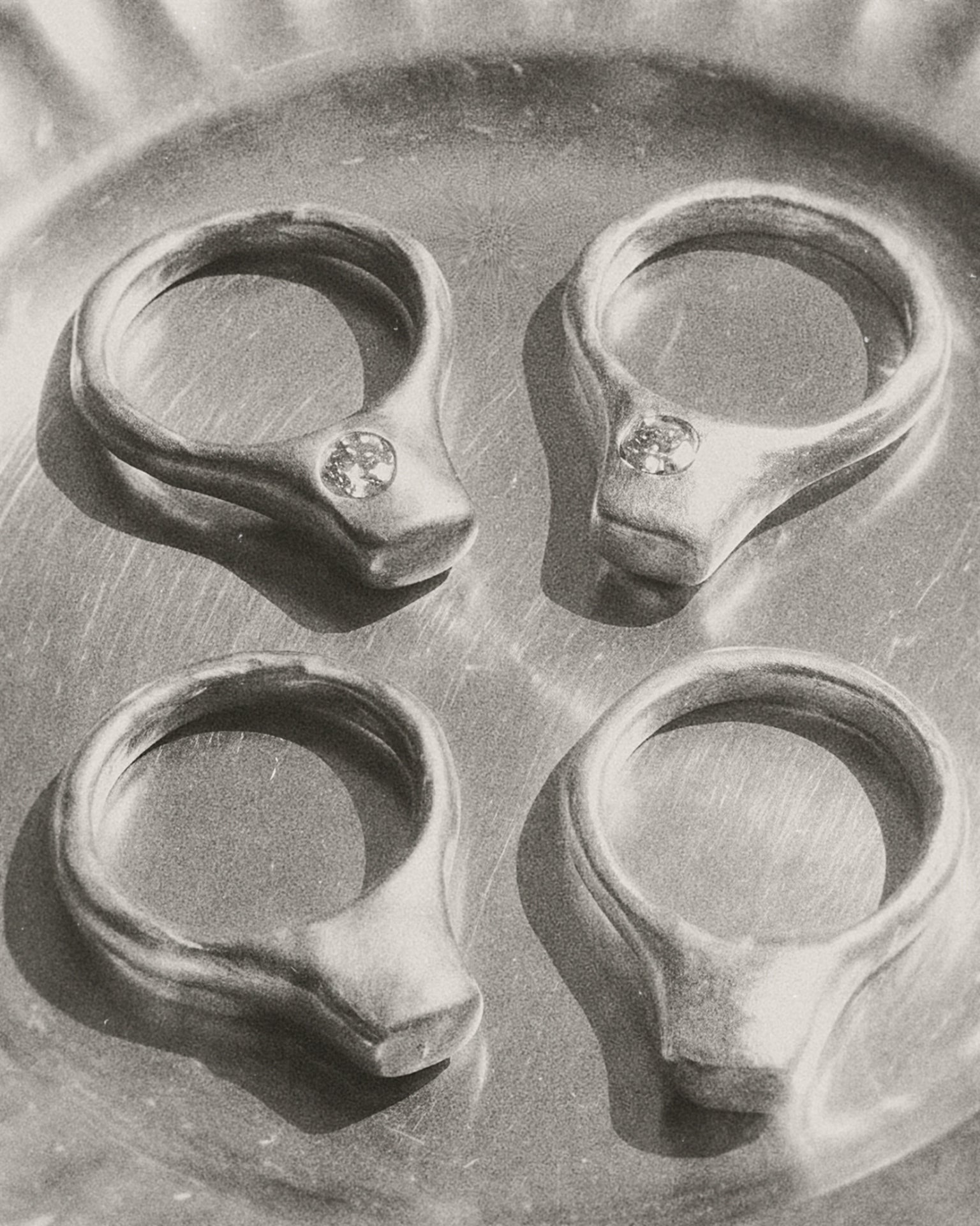 Four silver-toned signet-style rings with small rdiamond set in off-center, arranged in a 2x2 grid on a metallic surface.