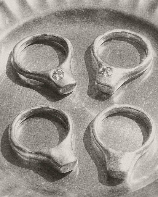 Four silver-toned signet-style rings with small rdiamond set in off-center, arranged in a 2x2 grid on a metallic surface.