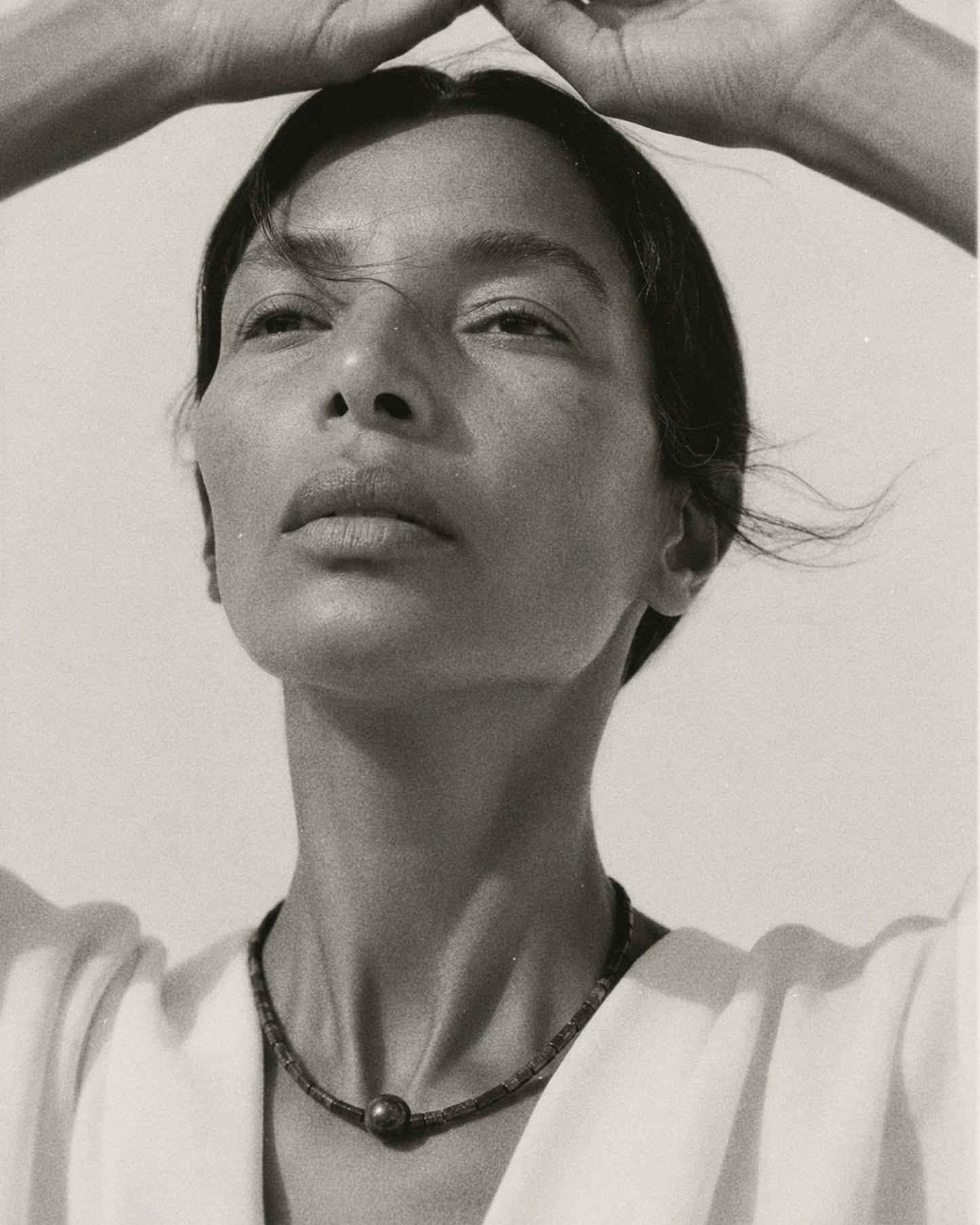 Black and white portrait of a woman looking upward with her hands forming a heart shape above her head, wearing a dark beaded necklace and a light top.