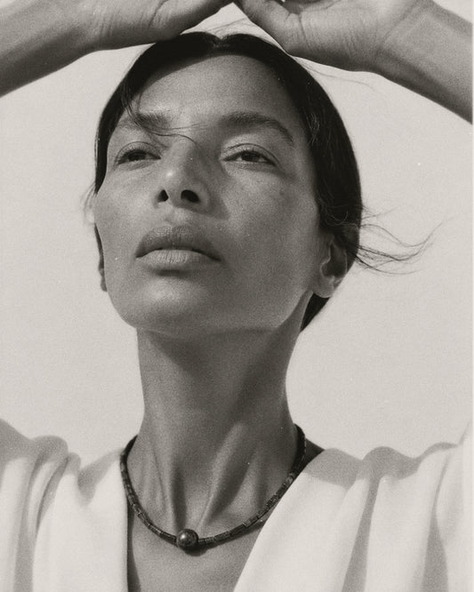 Black and white portrait of a woman looking upward with her hands forming a heart shape above her head, wearing a dark beaded necklace and a light top.