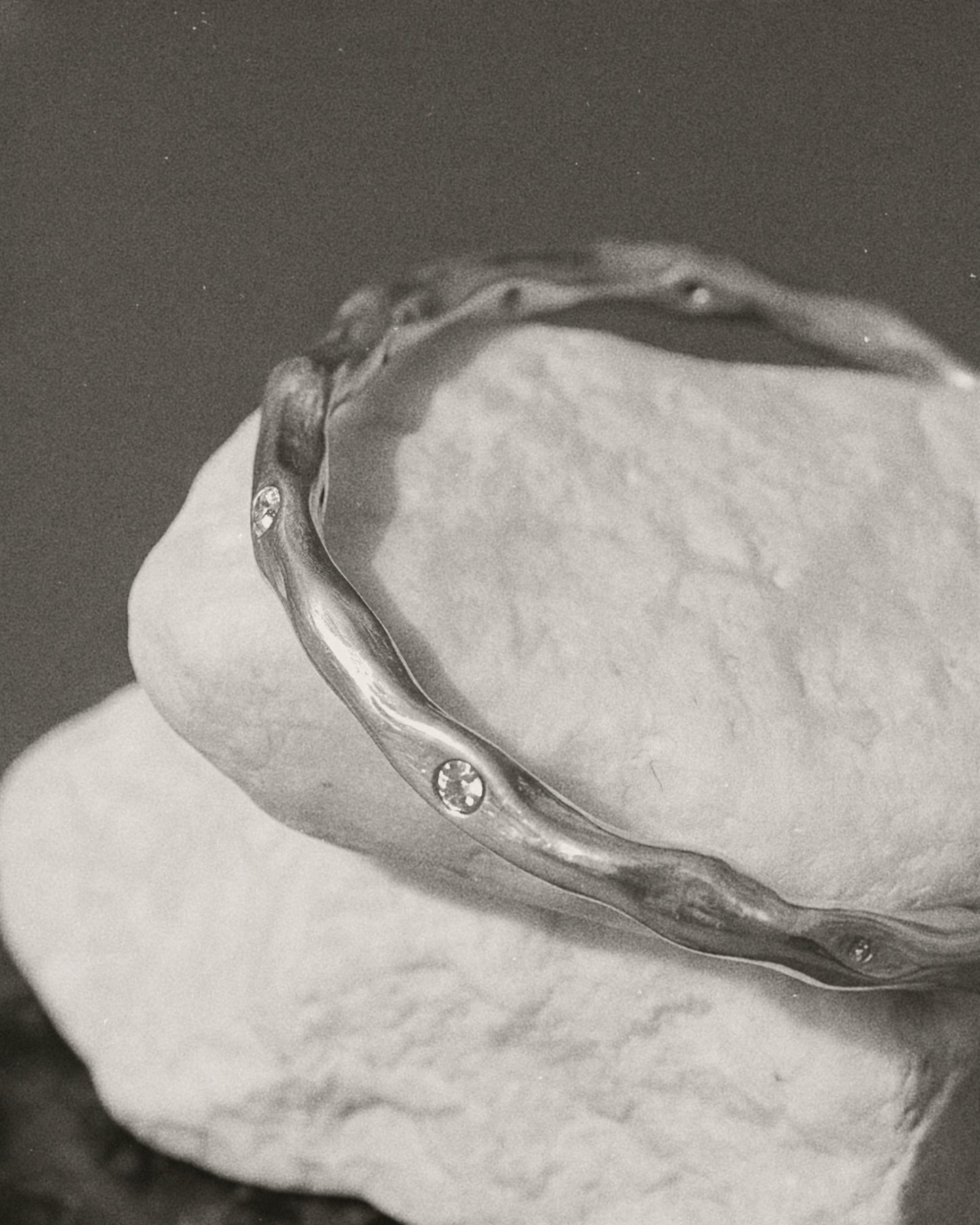 A silver-toned wavy bangle bracelet with small inset stones, displayed on a textured white stone against a dark gray background.
