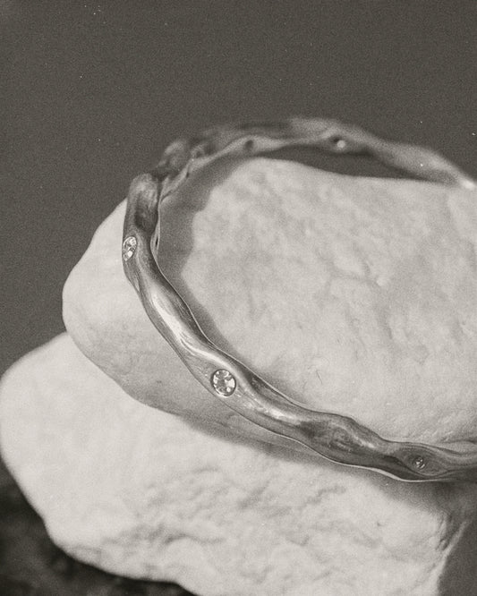 A silver-toned wavy bangle bracelet with small inset stones, displayed on a textured white stone against a dark gray background.