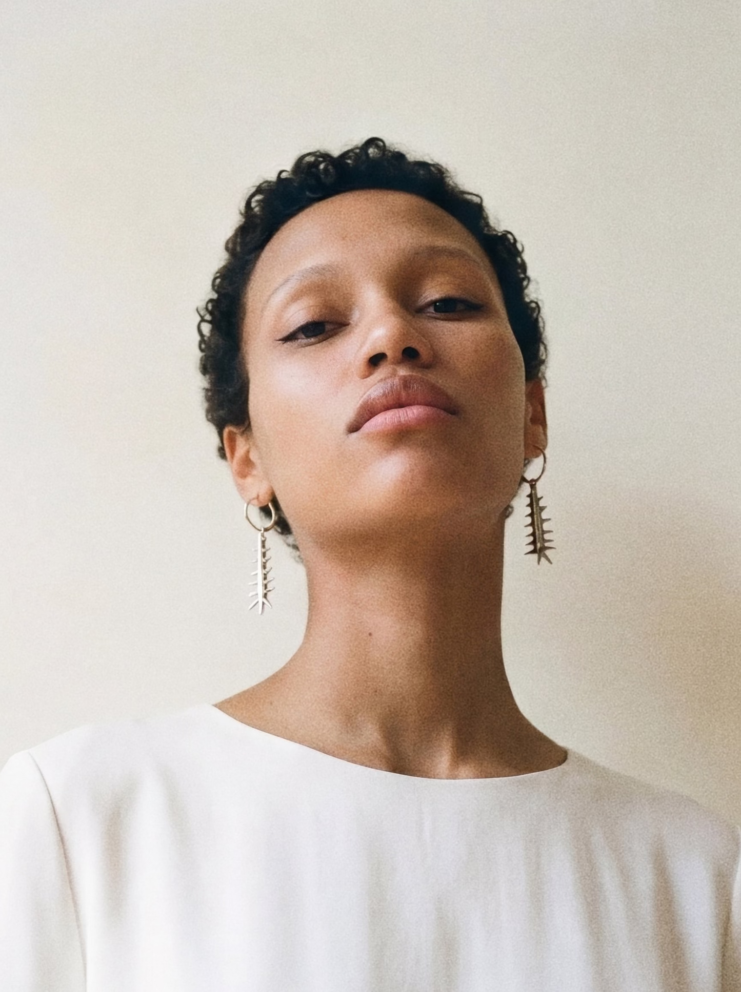 Woman wearing a white top against a plain background. She is wearing spiked earrings.